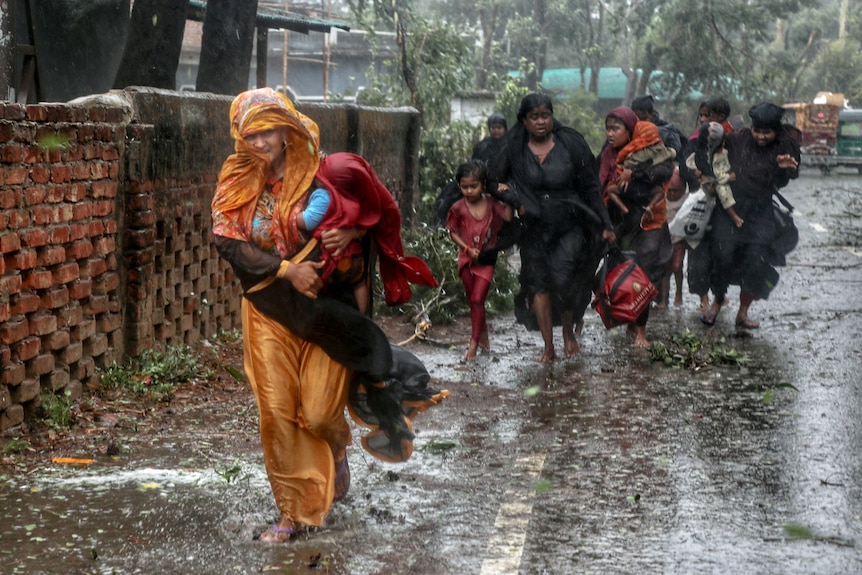 Women hold children and walk on flooded streets while drenched by rain.