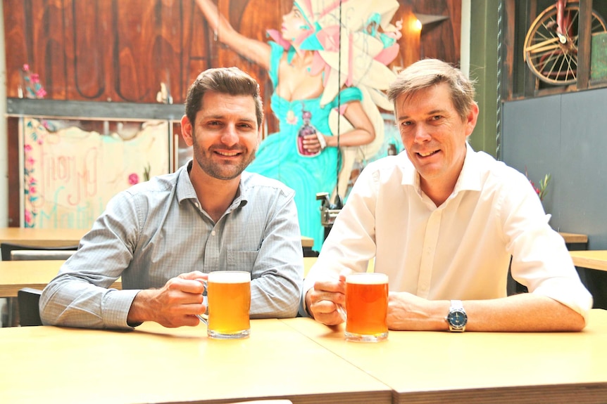 Two men sitting in a pub with beer glasses in hands and a mural in background
