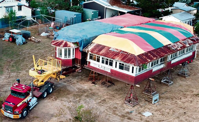 An aerial shot of a large truck driving away from a house on stilts with colourful tarps on the roof