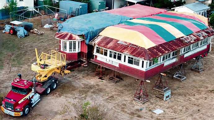 An aerial shot of a large truck driving away from a house on stilts with colourful tarps on the roof