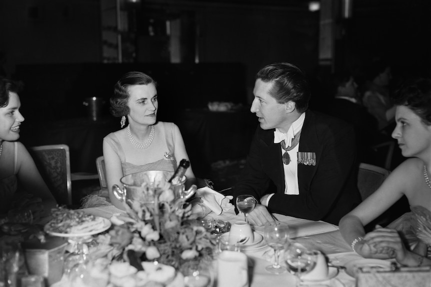 A woman sits a table in a dress listening to a man dressed in a dark suit.