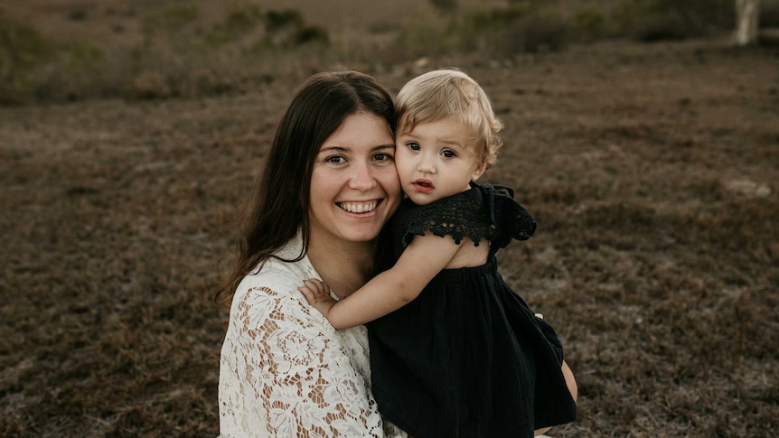 A young woman holds her toddler and smiles at the camera.