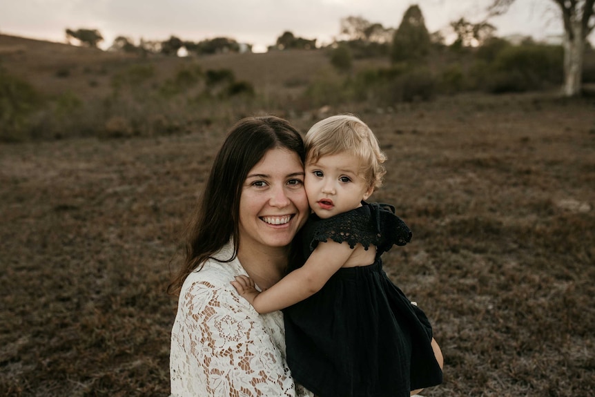 A young woman holds her toddler and smiles at the camera.