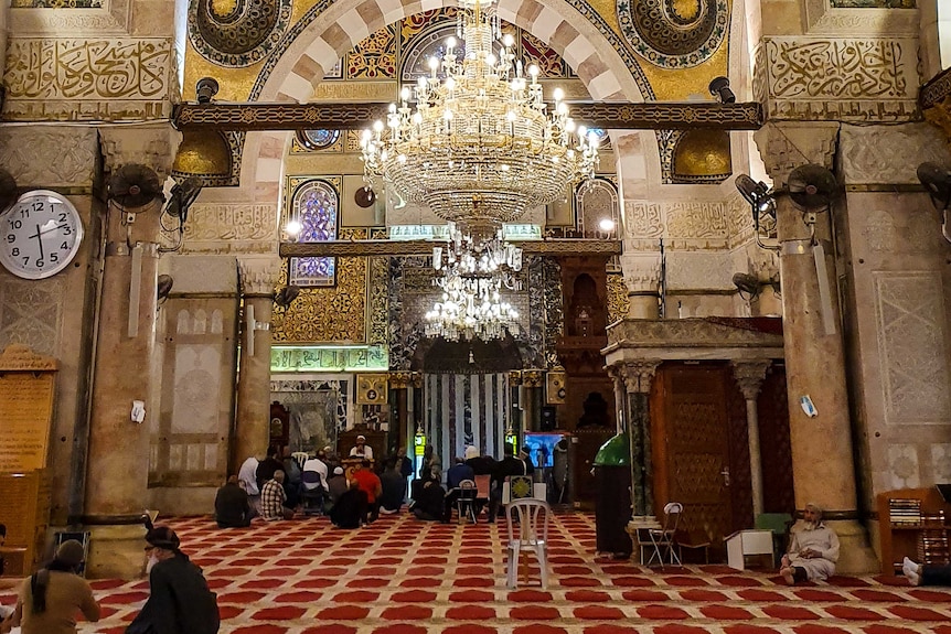 A room with red carpet and huge chandelier and people sit on the floor