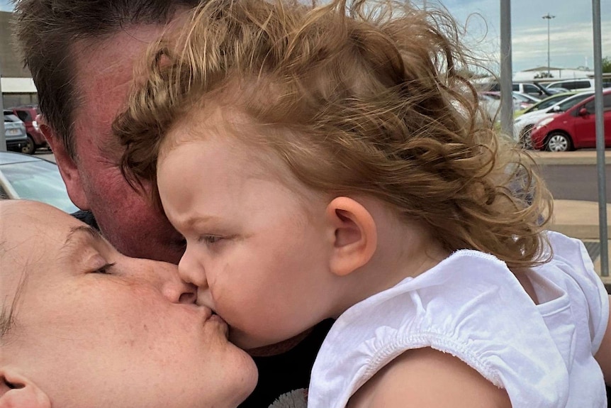 A mum kissing a two-year-old girl as a dad holds the girl. Cloudy day. in a carpark.