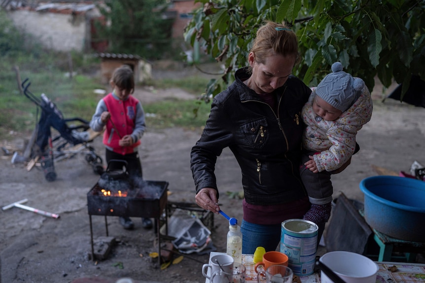 A woman holding a baby puts formula into a bottle while a child behind her puts a stick into a fire. 