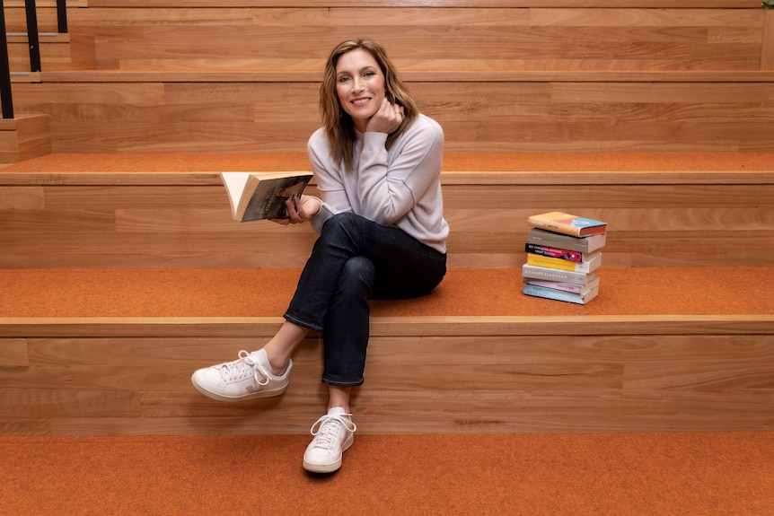 A woman sits on wooden stairs holding an open book and with a stack of books next to her. 