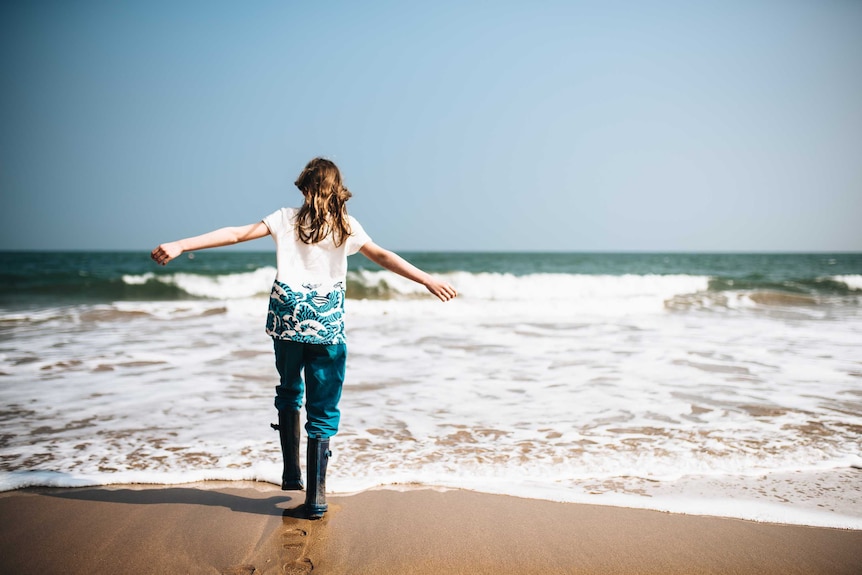 A woman facing the ocean holding her arms out with no-one else around for a story about taking mum vacations alone.