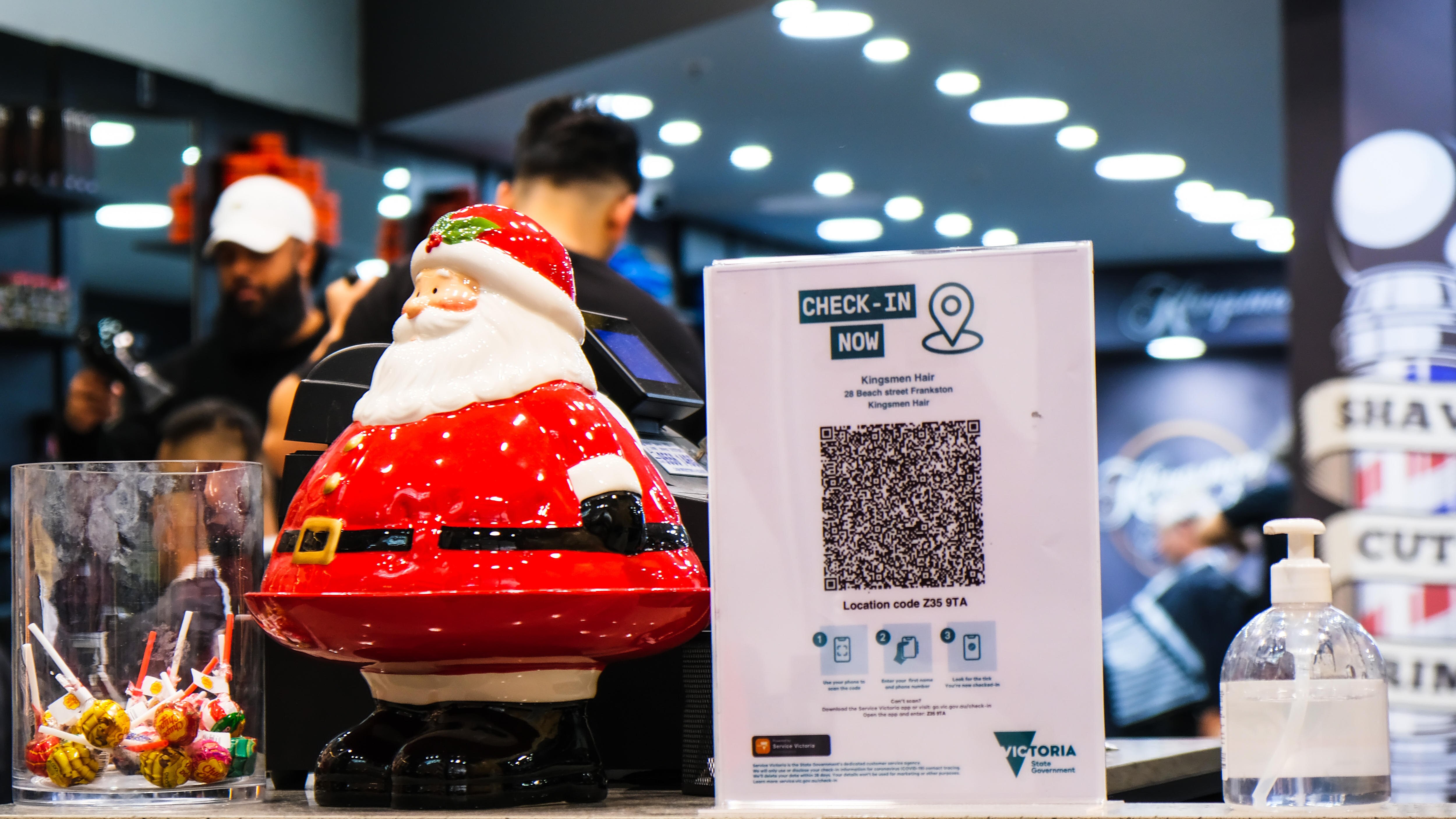 A tiny santa sits on a shop counter next to a QR check in board and a hand sanitizer and a jar of lollies.