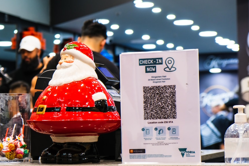 A tiny santa sits on a shop counter next to a QR check in board and a hand sanitizer and a jar of lollies.