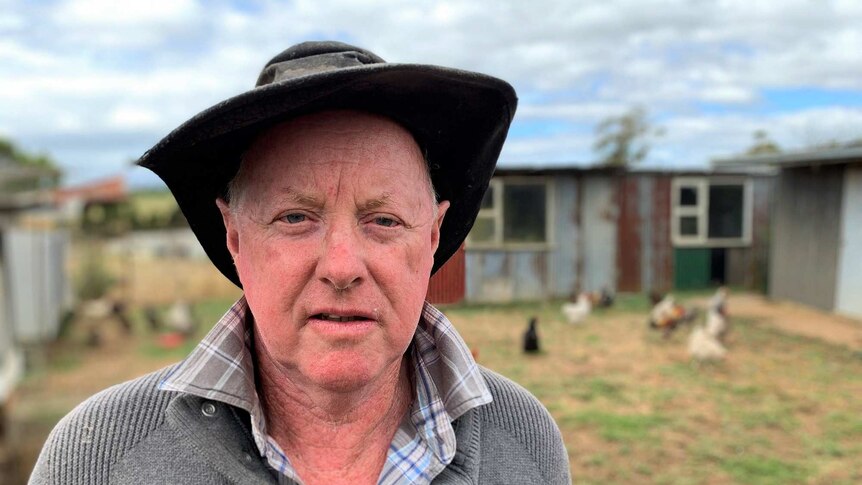 An older man wearing an hat stands in front of a shed and looks at the camera.