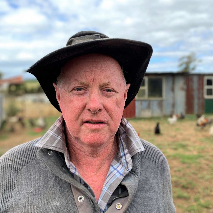 An older man wearing an hat stands in front of a shed and looks at the camera.