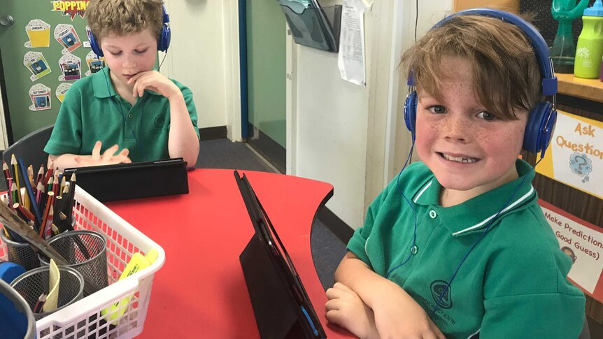 One student wearing headphones looks down at an iPad while another wearing headphones and sitting in front of an iPad smiles.