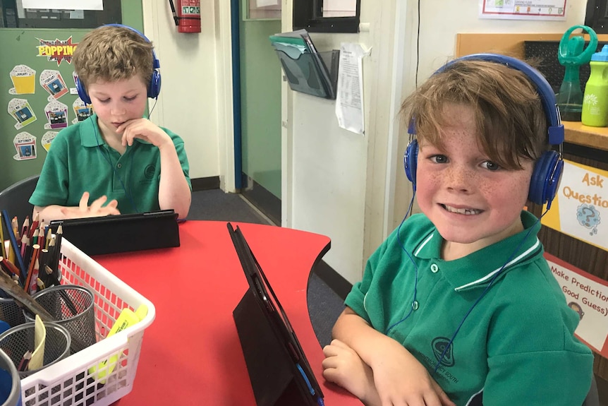 One student wearing headphones looks down at an iPad while another wearing headphones and sitting in front of an iPad smiles.