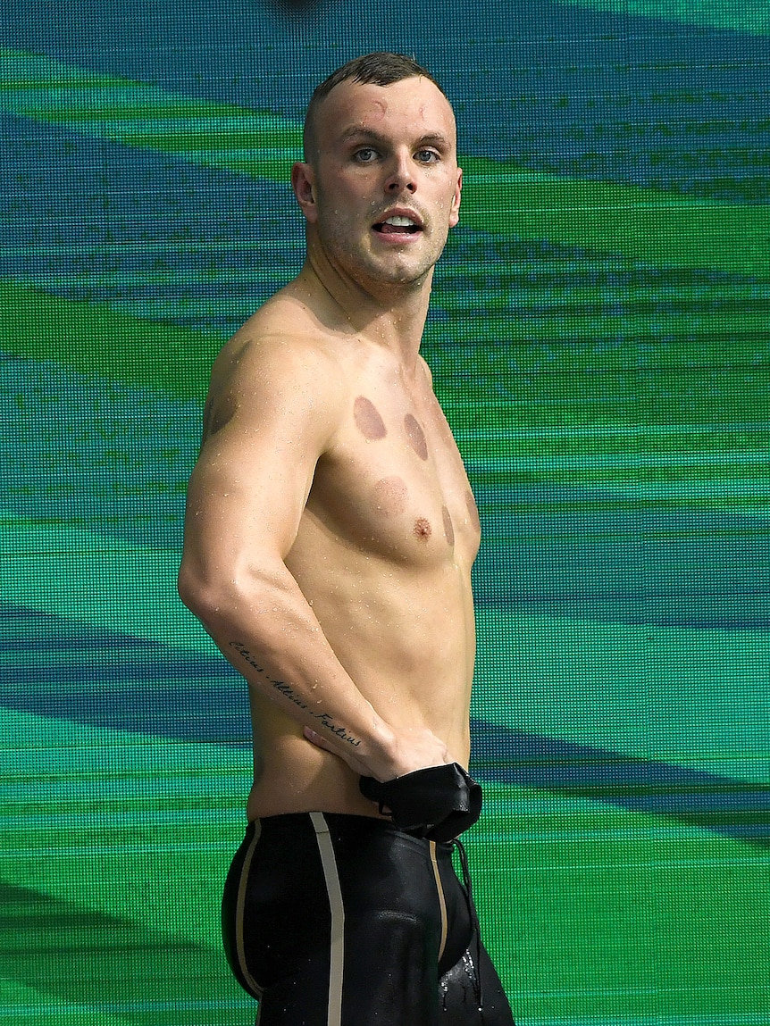 A man in swimming trunks stands with his hands on his hips.