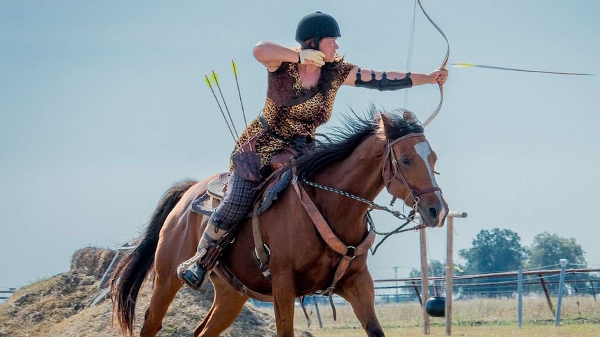A woman wearing leopard print and a riding helmet about to shoot an arrow while sitting on a horse.