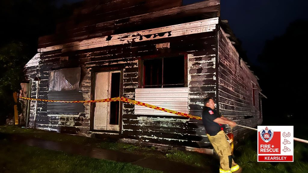 A burnt out weatherboard house. Firefighter ties yellow and red tape across the front of it.