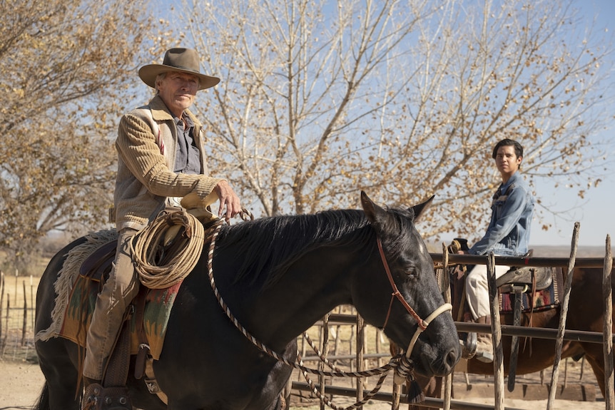 A 90-something man astride a black horse and a Hispanic teenager on a brown horse look concerned