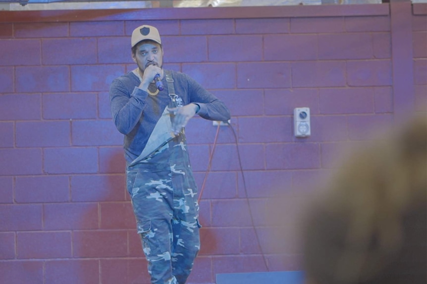 Wayne 'Dubb T' Tobane wearing a cap, overalls, long sleeve shirt, rapping, holding a microphone, brick wall behind.