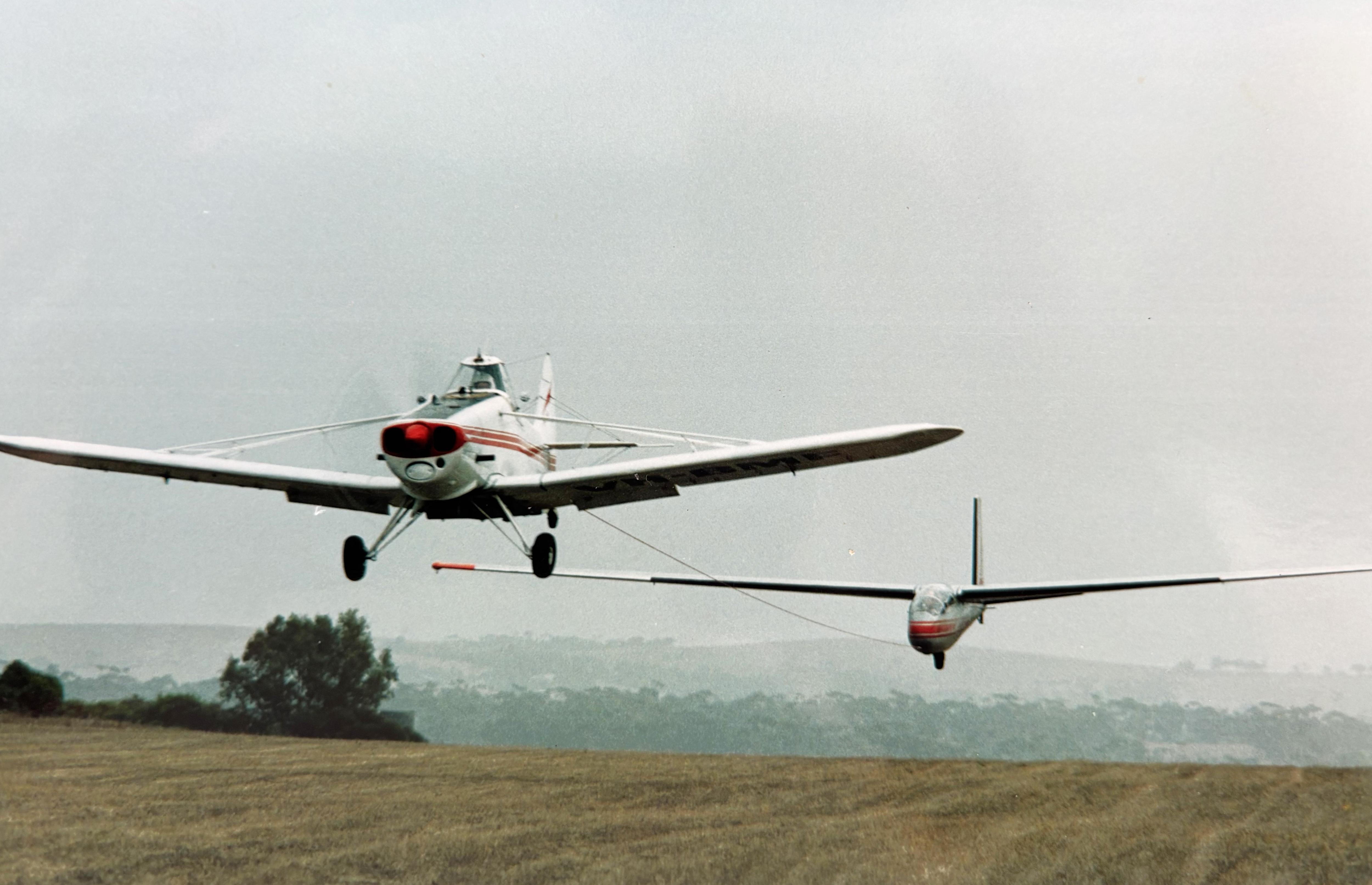 Single cabin white plane towing a glider, only about five metres off the ground, over a paddock.