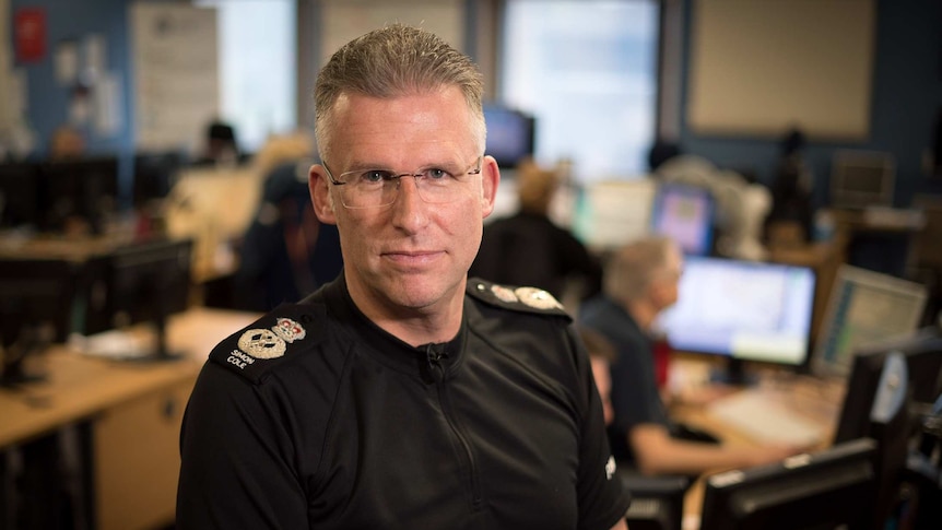 In a dim office, a Caucasian police officer in black looks up to camera as his staff work on computers blurred behind him.