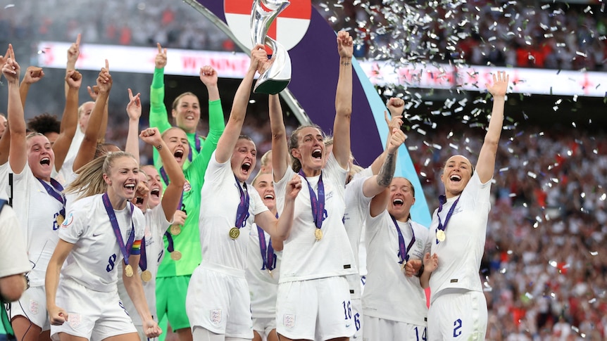 England's women's soccer team hold a trophy in the air and pump their arms in the air in celebration. 