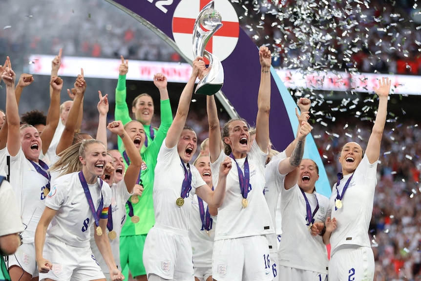 England's women's soccer team hold a trophy in the air and pump their arms in the air in celebration. 