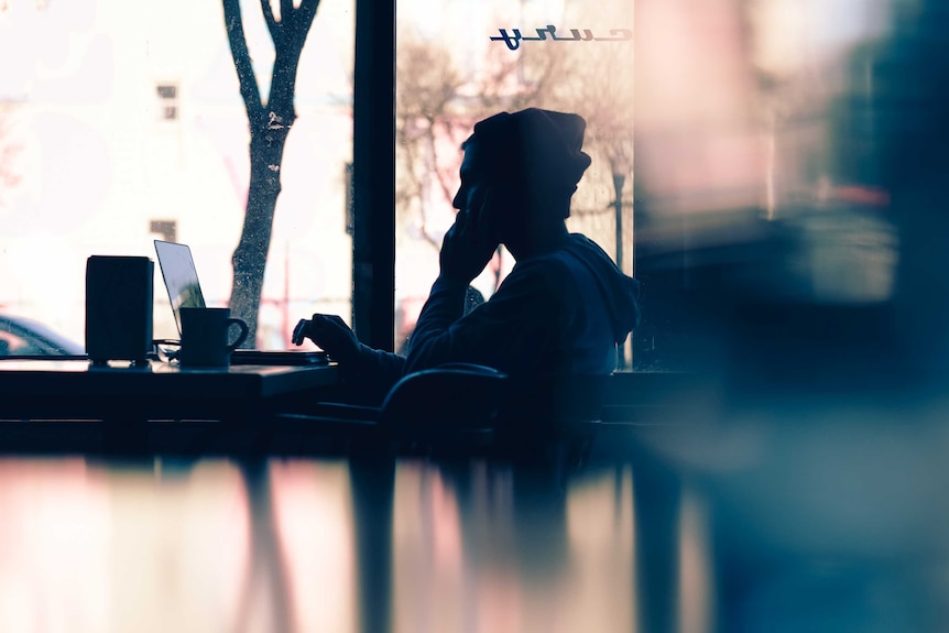 A man looks at a laptop silhouetted against a cafe window.