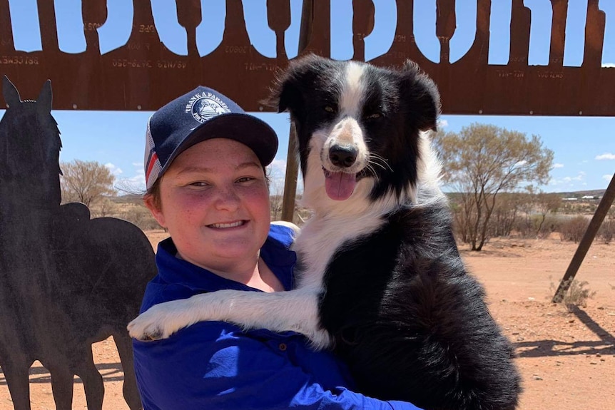 A young woman in a hat holds a black and white dog in front of a sign.