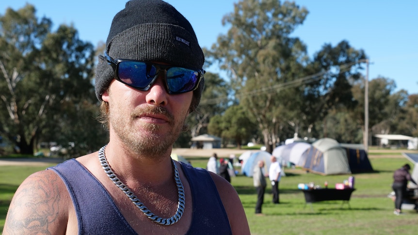 A man wearing a beanie, sunglasses and a silver chain stands in front of a number of tents at a campsite.