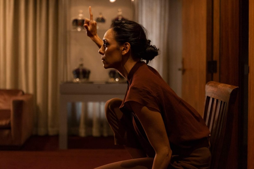 Side profile shot of a woman dressed in brown sitting on a chair, interior, with her right arm raised and forefinger pointed up.