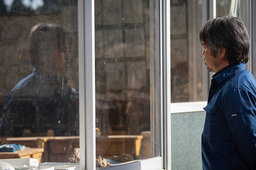 A Japanese man in a blue shirt looks through the window of an abandoned classroom