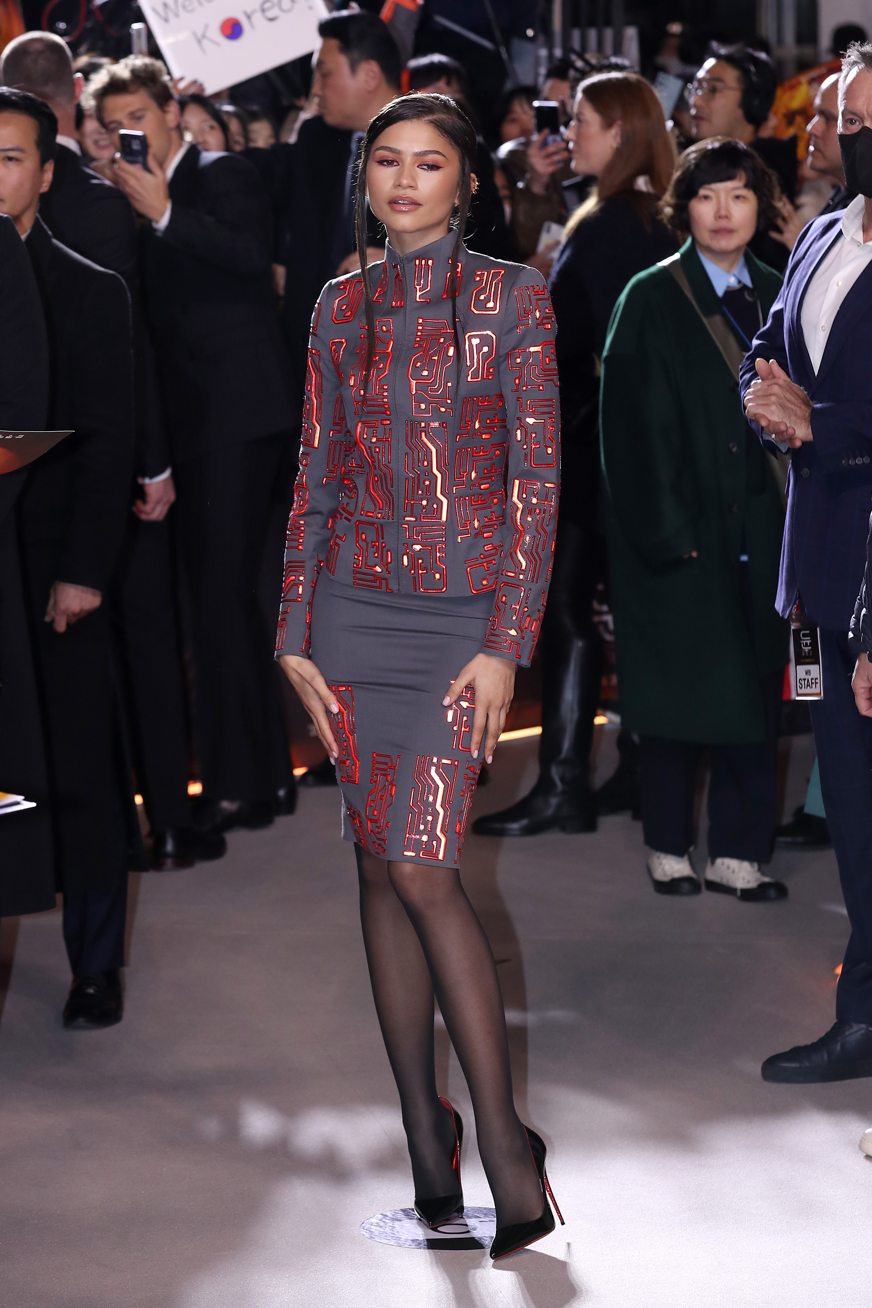Zendaya poses in a long-sleeved grey top and matching pencil skirt that's covered in red sequins that look like computer coding