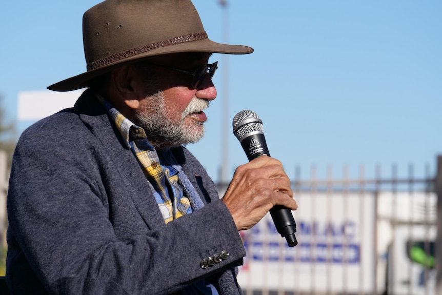 Harold Furber, an Indigenous elder, holds a microphone. He is wearing a brown hat and a blue jacket, with a checked shirt.