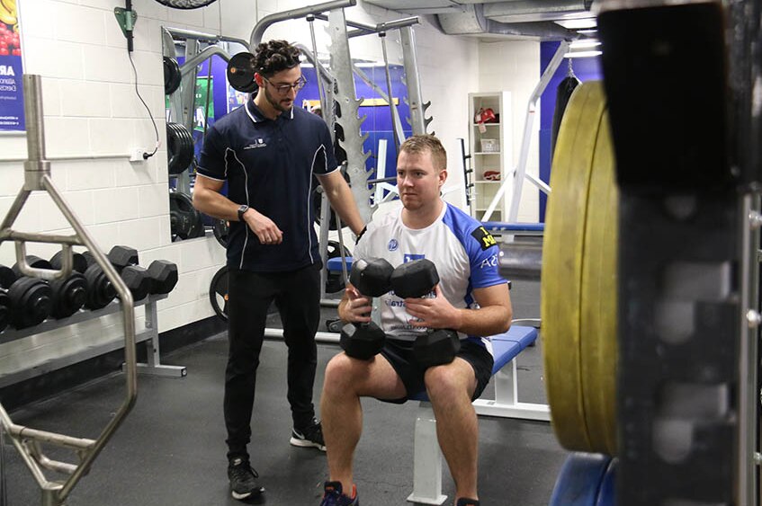 A man sits on a weight bench holding 20kg dumbbells while a trainer observes him