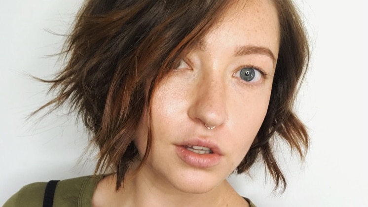 A close-shot of a woman with short wavy hair and dark green top, and nose ring, with neutral expression.