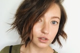A close-shot of a woman with short wavy hair and dark green top, and nose ring, with neutral expression.