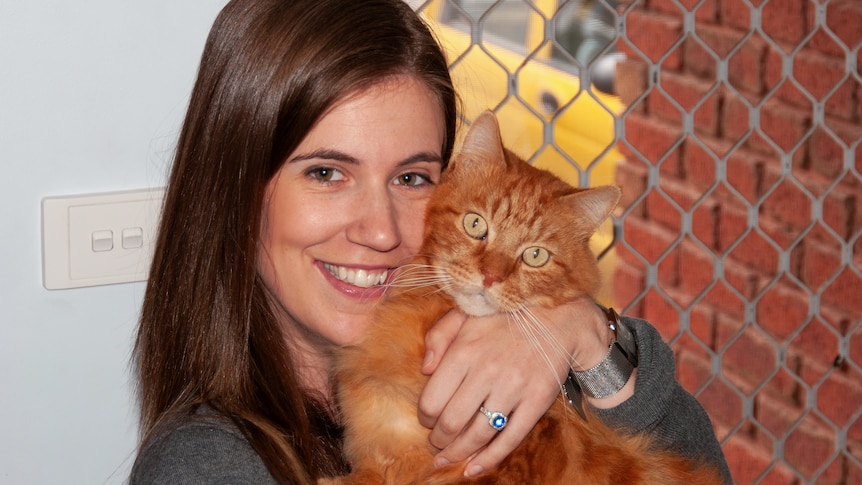 Smiling woman with brown hair cuddles a ginger cat, both look to the camera.