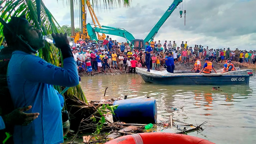 People crowd around a river bank watching officials in a boat. 