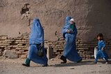 Two women in blue burkas walk across a dusty path. One carries a baby, a child dressed in blue follows.