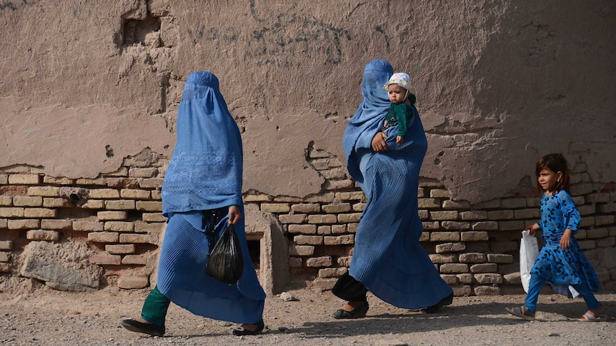 Two women in blue burkas walk across a dusty path. One carries a baby, a child dressed in blue follows.
