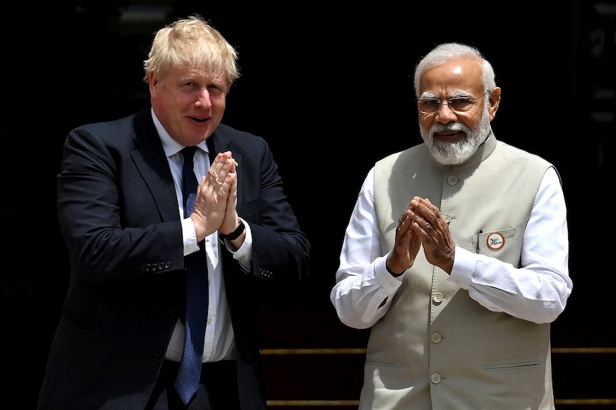 Johnson and Modi stand side by side, looking out towards the camera. They both have their palms pressed together in applause
