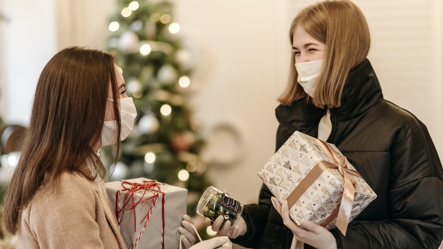 Two young women wearing face masks and holding wrapped Christmas presents while looking at each other