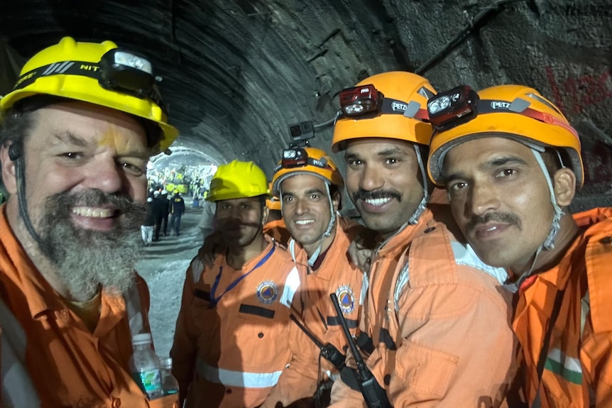 A group of miners in orange hi-vis gear and helmets smile inside a dark tunnel