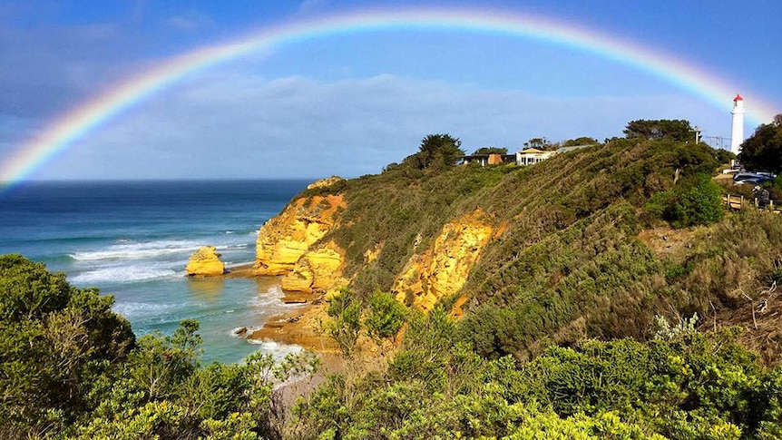 A rainbow stretches from the beach over the orange cliffs of aireys inlet to the famous local white with red roof lighthouse