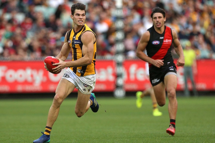 An AFL player runs hard with the ball through the midfield, chased by an opponent.