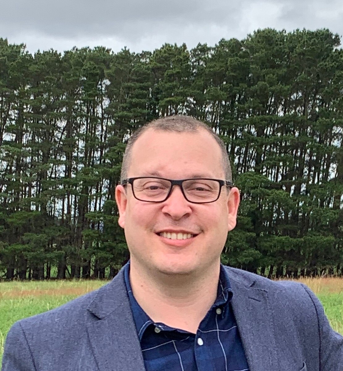 timber A man wearing glasses and a suit stands in front of pine trees.
