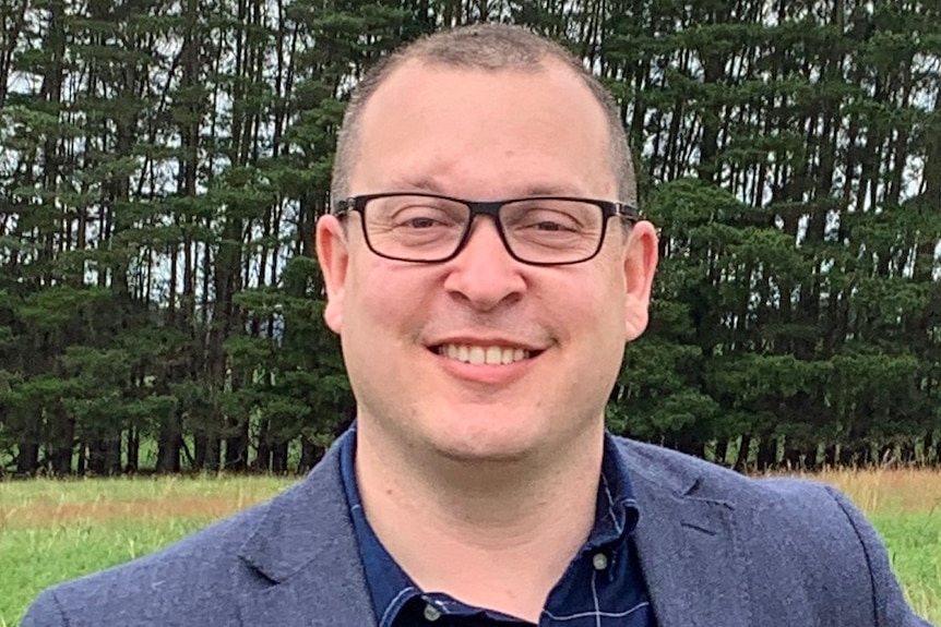 A man wearing glasses and a suit stands in front of pine trees.