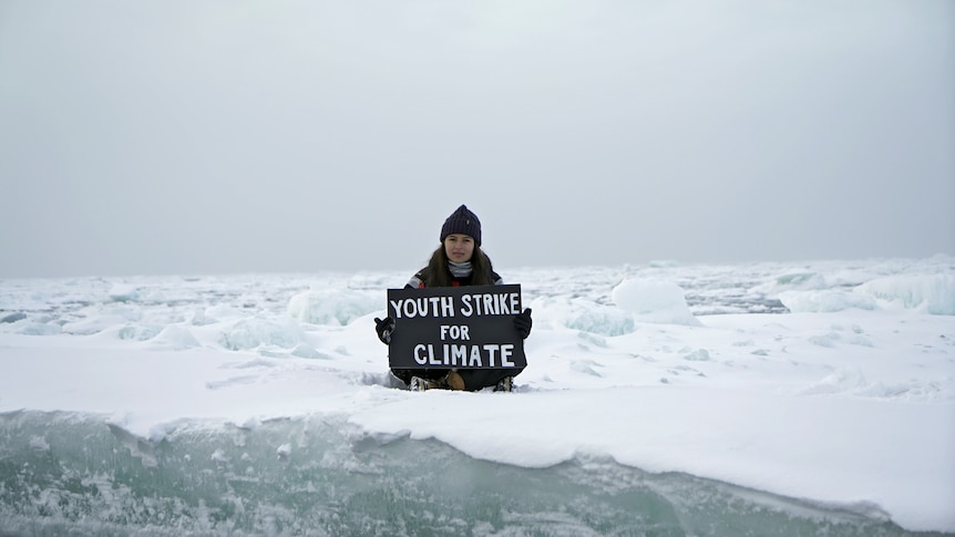 A girl dressed in dark snow clothes sitting crossed legged on the ice holding a sign saying "youth strike for climate".