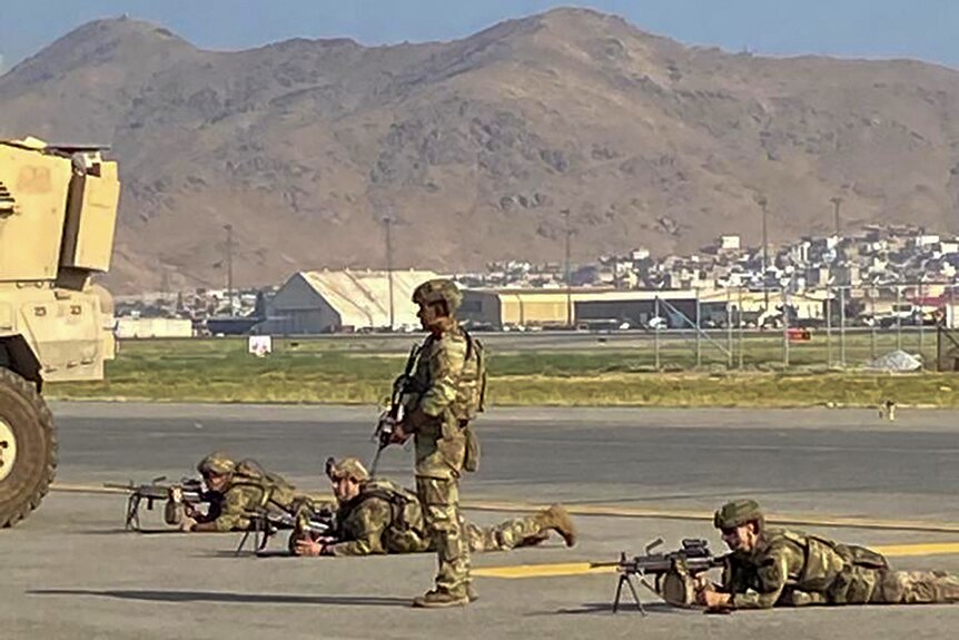 One armed soldier in military fatigues stands as three other lie down at airport flanked by a rugged hill.